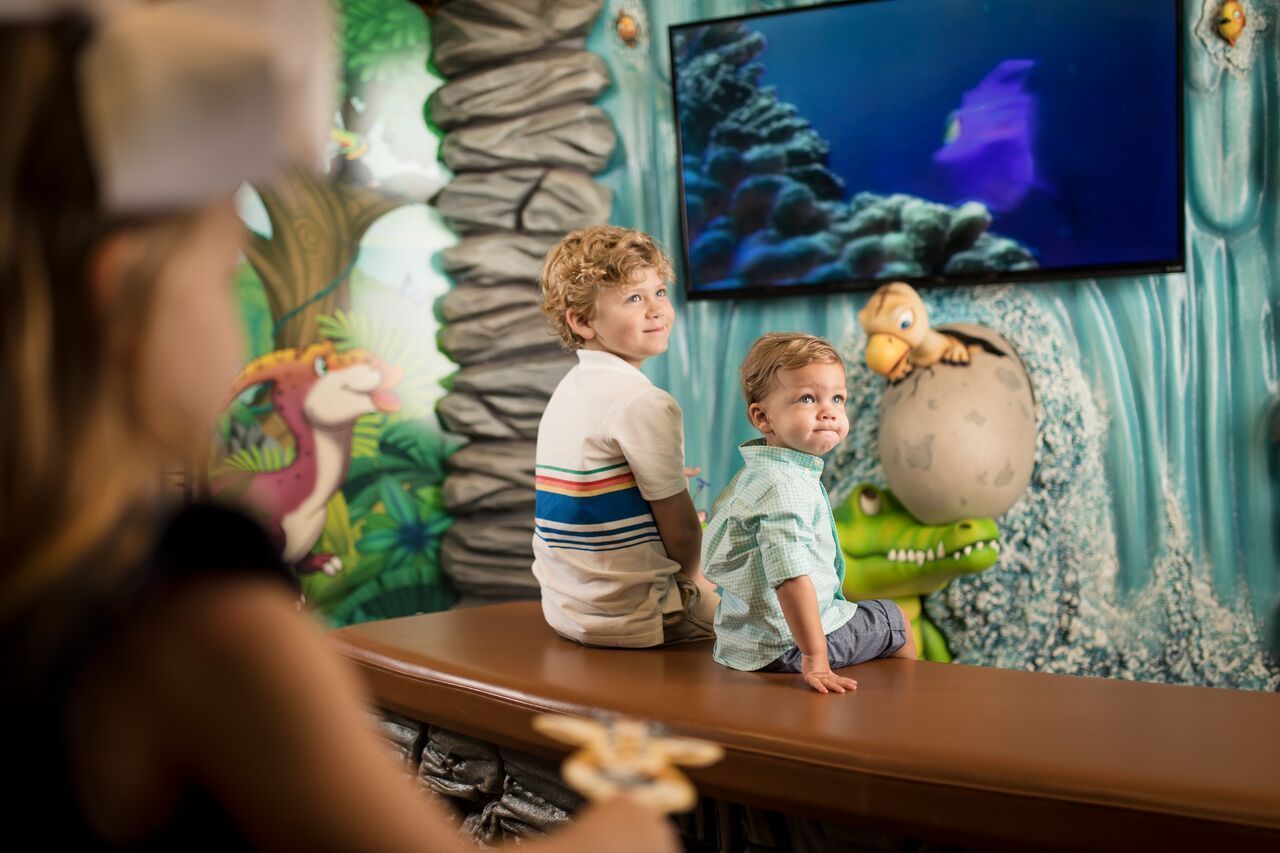 Two young boys watch a video in a kid-friendly room with dinosaur décor; one boy smiles, the other appears upset.