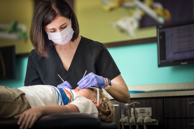 Dentist examining a young boy's teeth in a dental office; the dentist wears a mask and gloves.