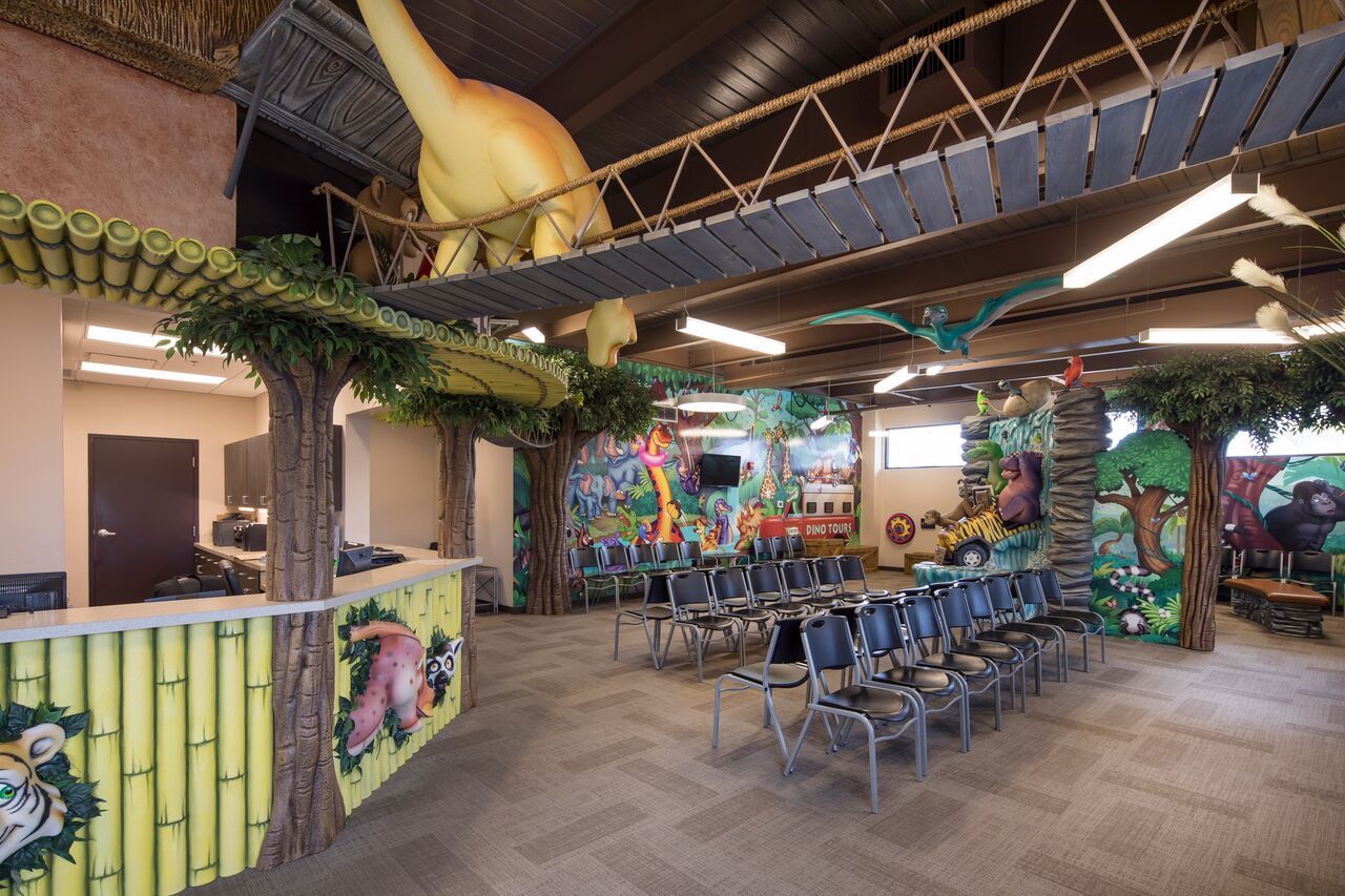 Dinosaur-themed waiting room. Giant dinosaur, bridge, jungle-themed walls, rows of chairs, and decorated columns.