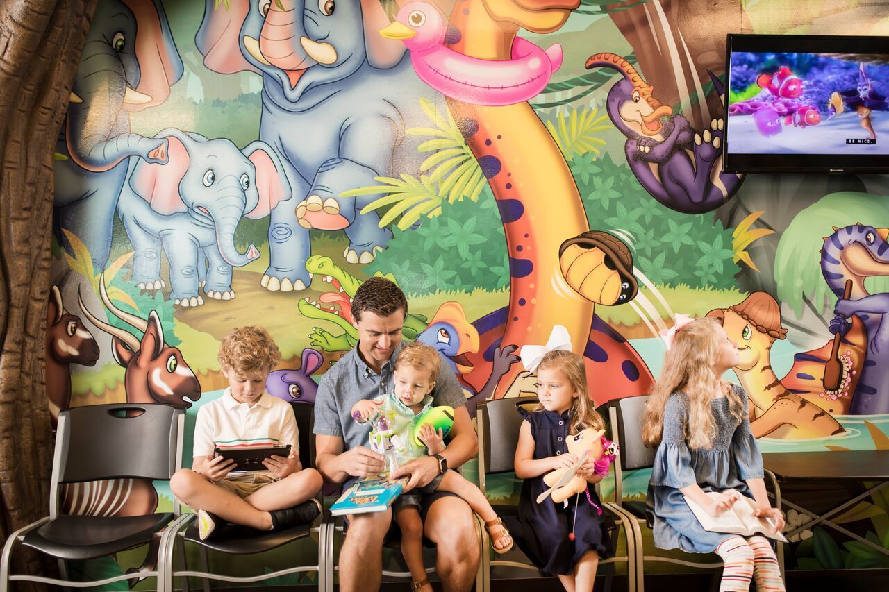People sitting in a dentist's office with jungle-themed murals. A man and three children are waiting.