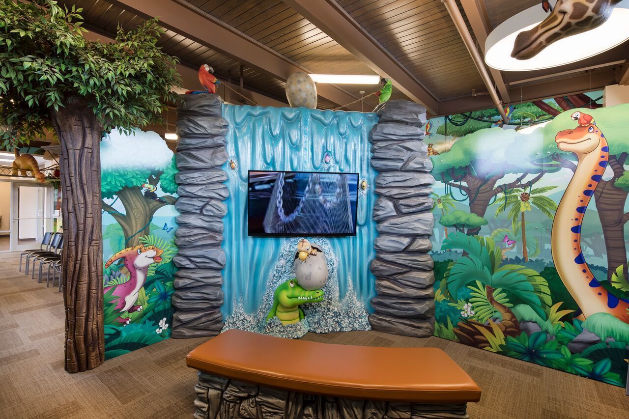 Waiting area in a pediatric dentist office with jungle mural, waterfall backdrop, bench, and TV.