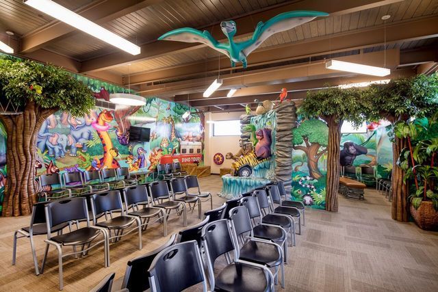 A dinosaur-themed kids' room with rows of chairs, murals, and a pterodactyl sculpture.