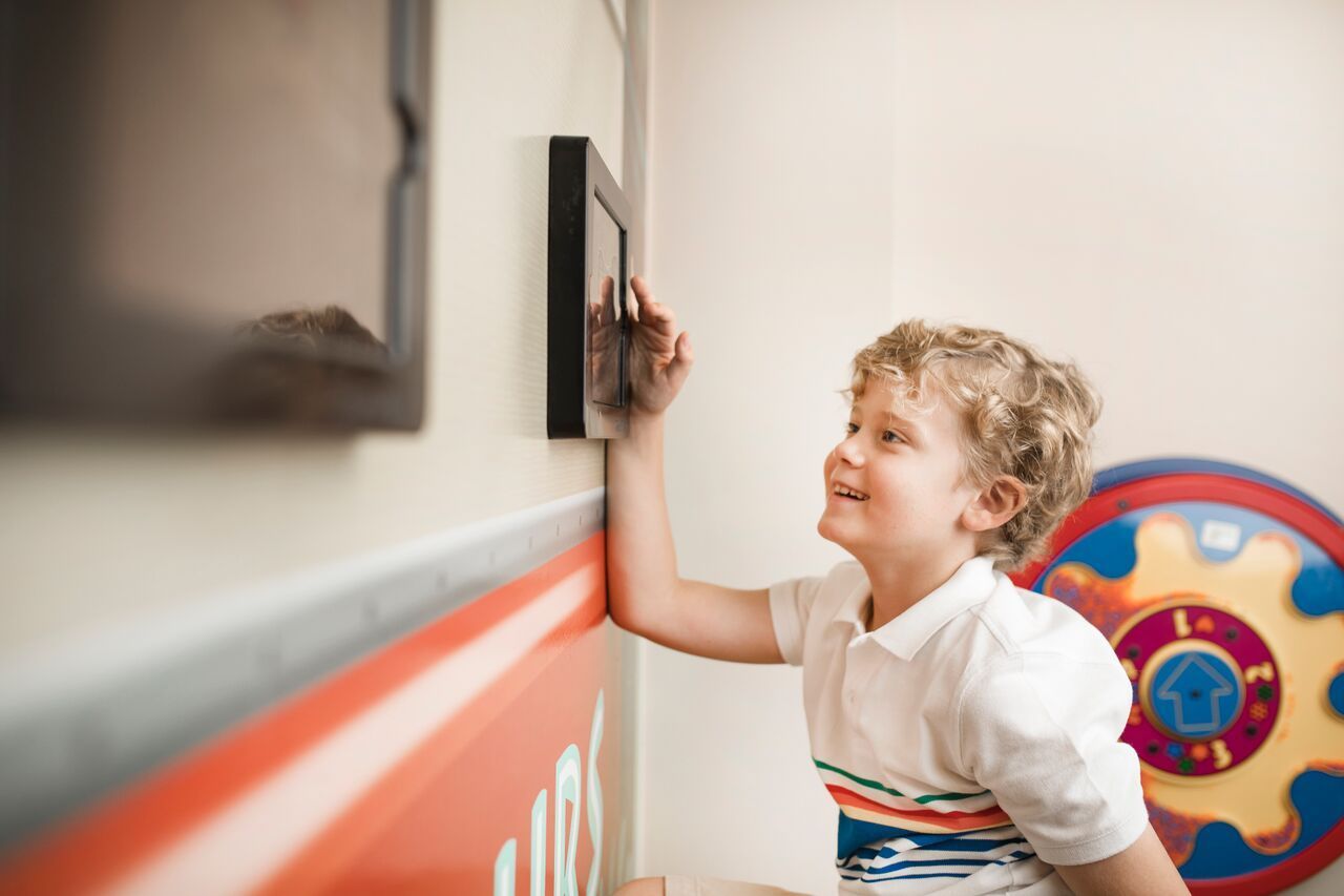 Boy touches a framed screen on a wall, smiling. Brightly colored toy on wall.