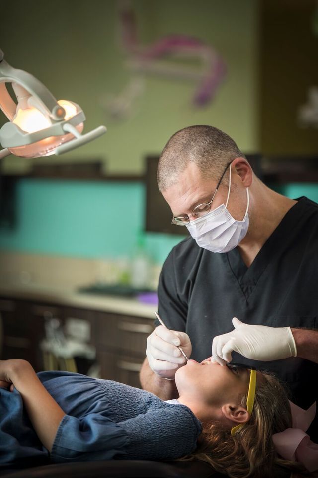 Dentist examining a patient's teeth. Man wearing mask and gloves; girl lying in chair; dental office interior.
