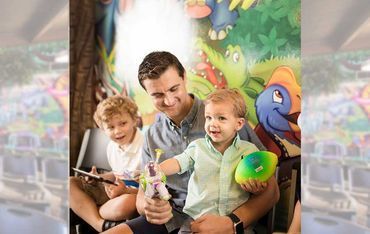Father smiling with two young sons, holding toys, in a colorful jungle-themed room.