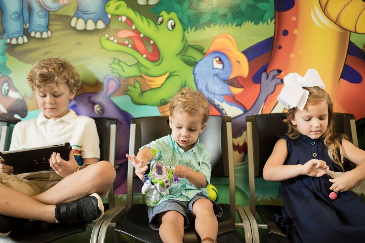 Three children in a waiting room, focused on a tablet, toy, and hands, with a dinosaur mural background.