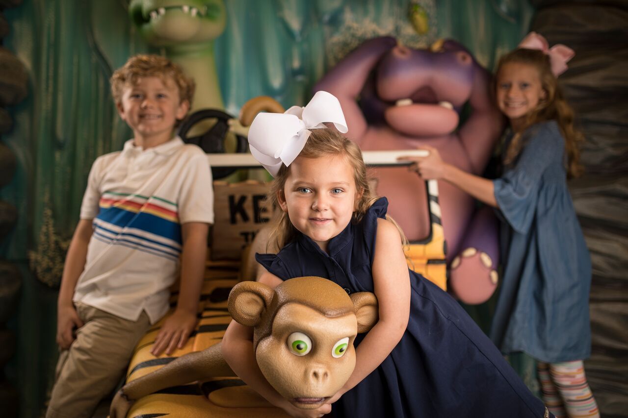 Three children pose with cartoon animal figures in a jungle setting.