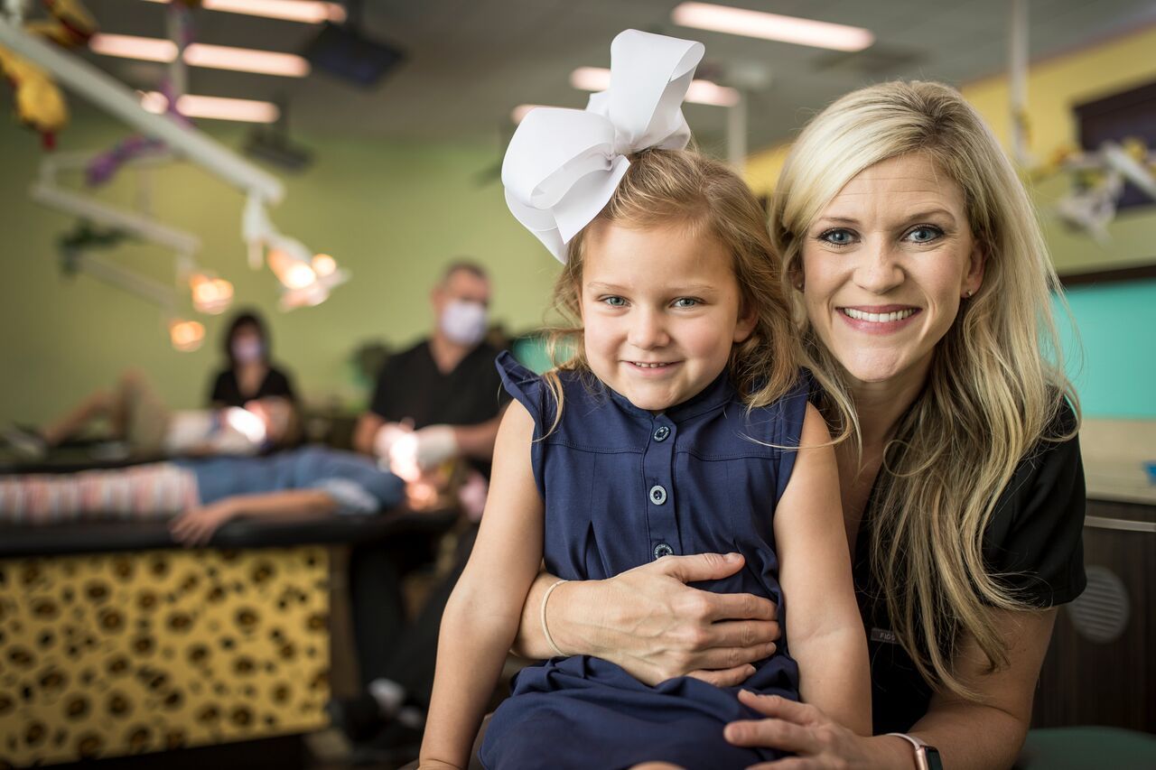 Woman and young girl smiling in a pediatric dentist office, while a dentist is treating a patient in the background.