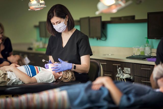 Dentist examining a child's teeth in an office setting; others await treatment.
