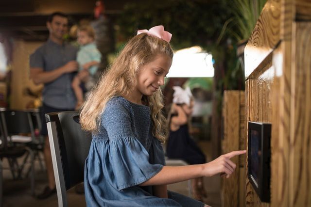 Girl with pink bow touches screen at a wooden booth. Family in background.