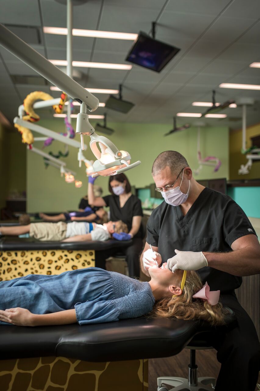 Dentist examining a girl's teeth with assistance in a brightly-decorated office. Other patients and a dental assistant are present.