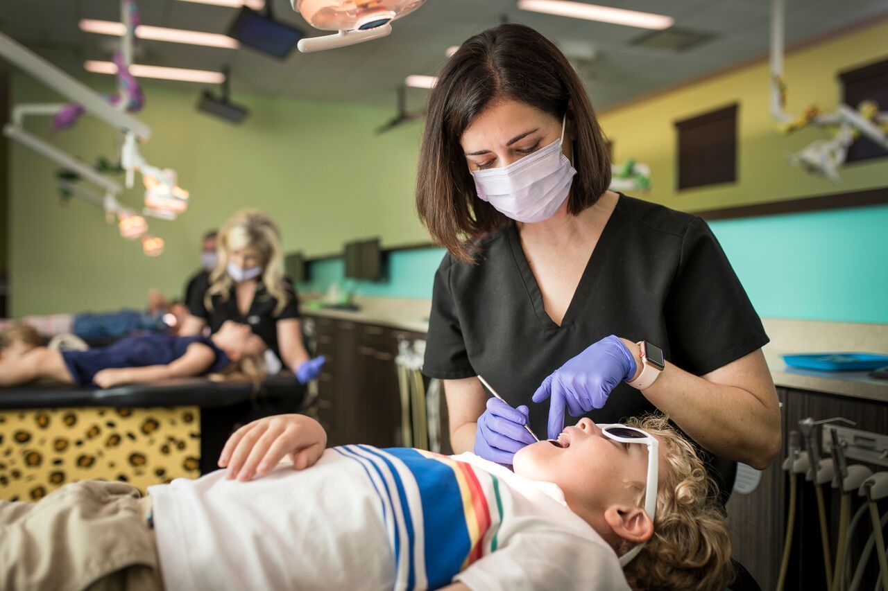 Dentist examining child's teeth in office. Both wearing masks. Other children and dentist in background.