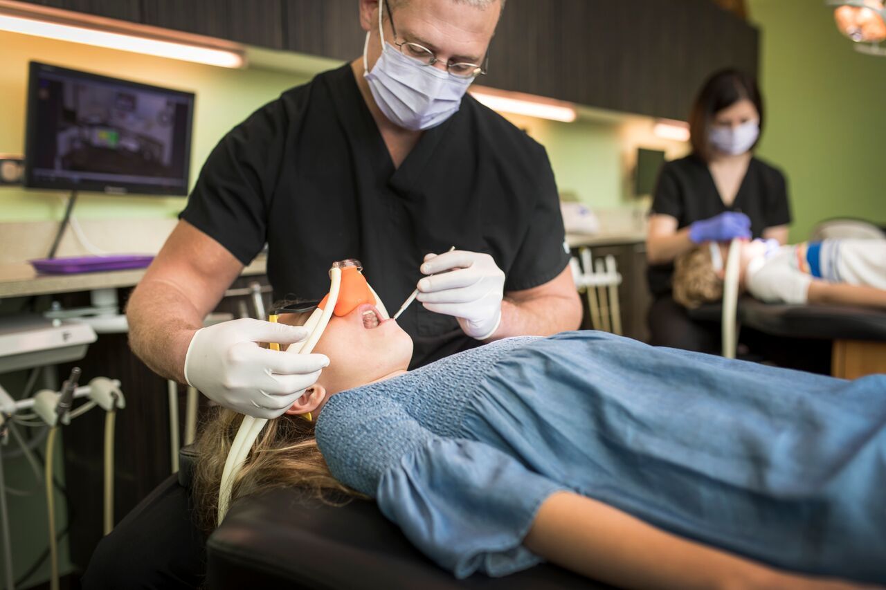 Dentist examining a patient's teeth. Assistant in background. Dental office setting.