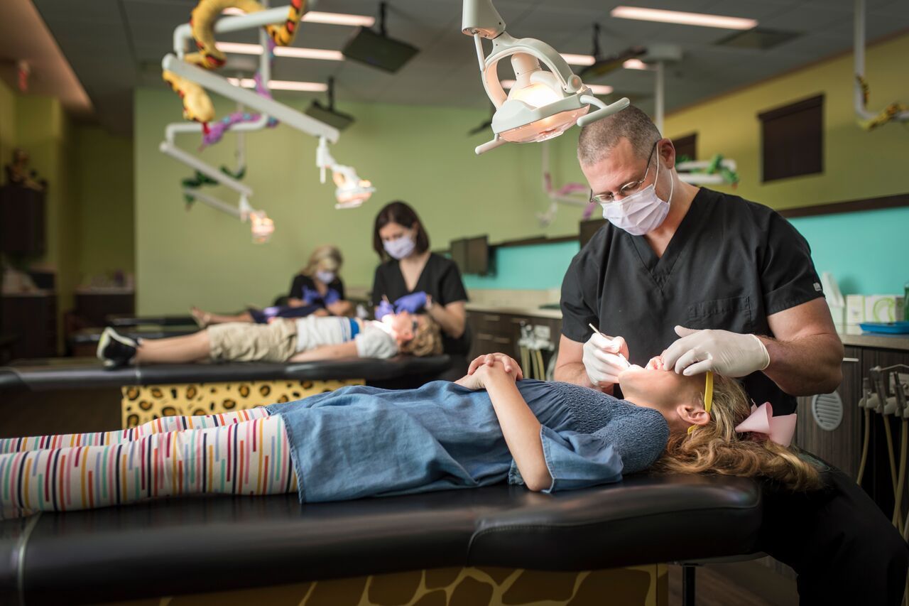 Dentist examining a young girl's teeth; children and staff in a colorful dental office.