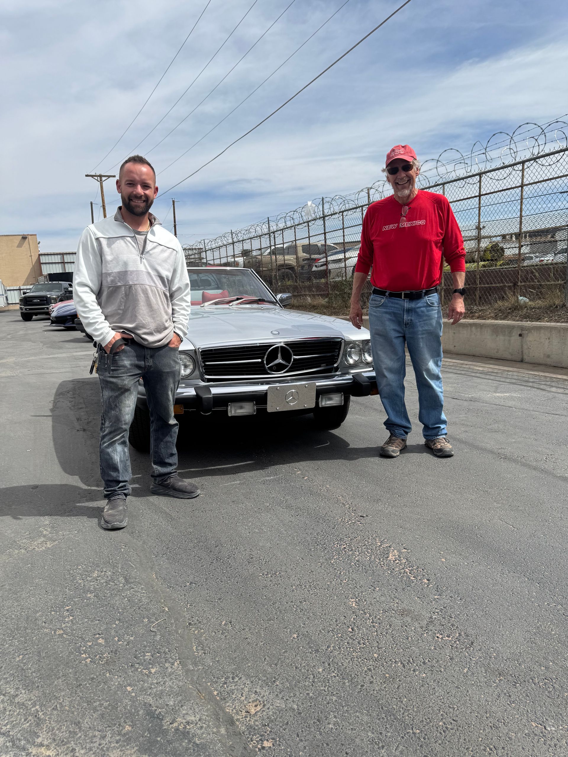 Two men standing beside a damaged car on a gravel lot under a cloudy sky.