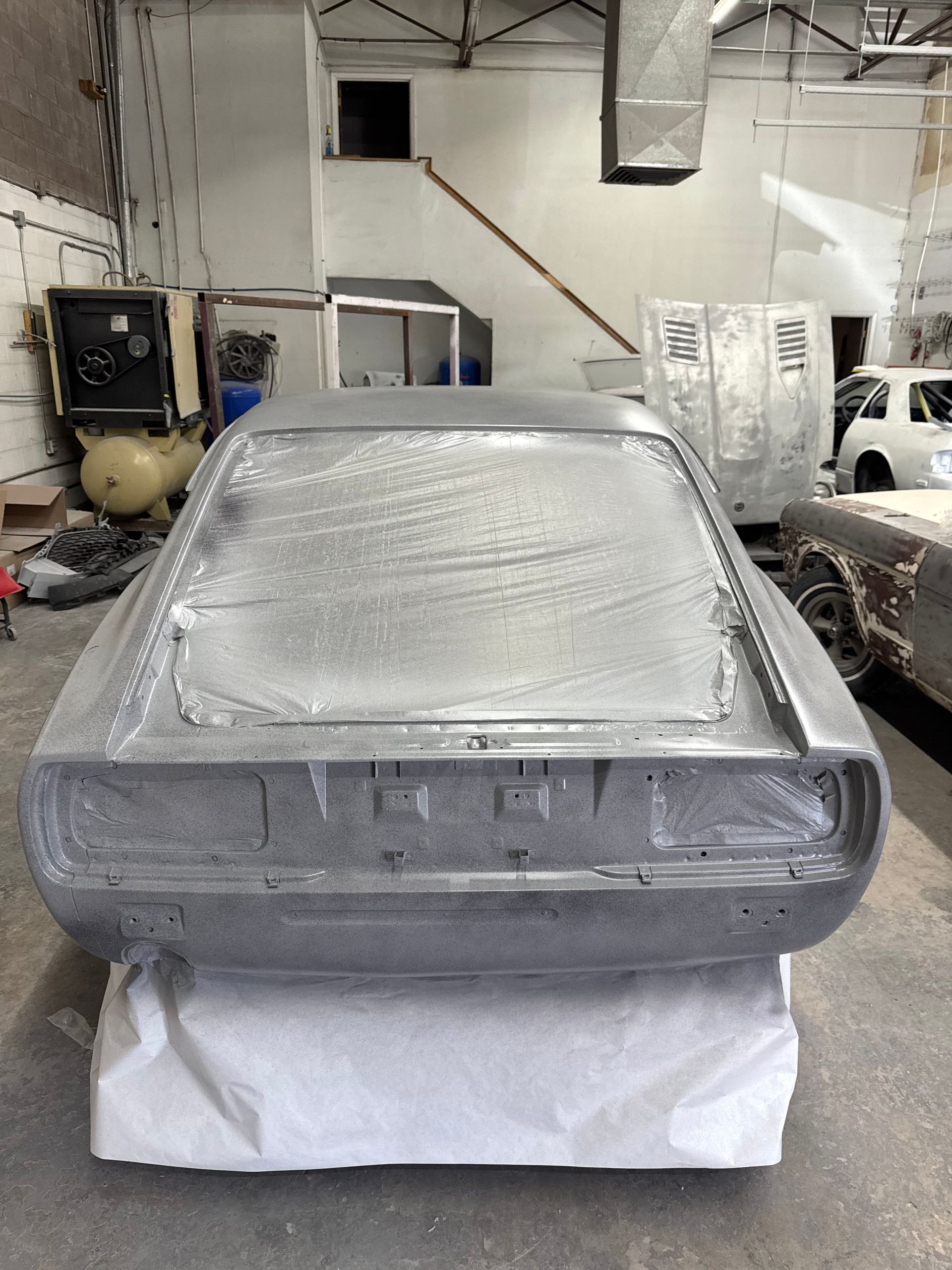 Metallic car body panel wrapped in protective plastic in a workshop, resting on a covered stand.