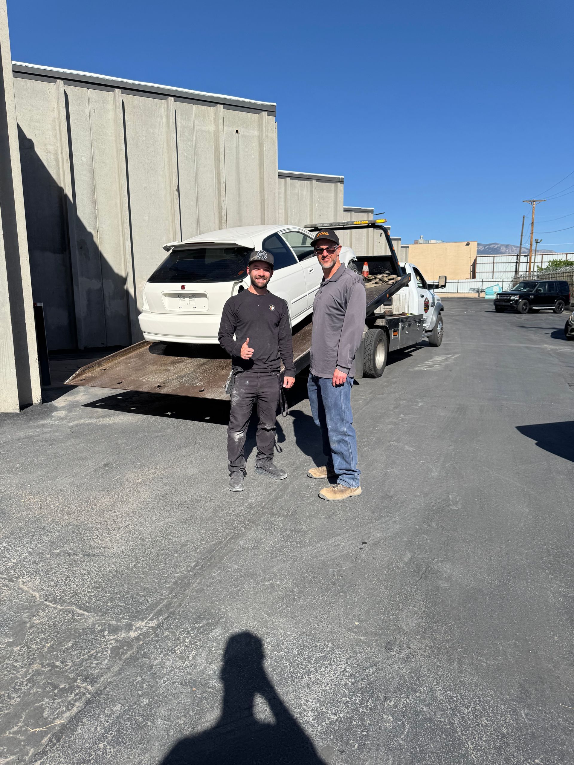 Two people standing by a white car in an industrial parking lot on a sunny day.