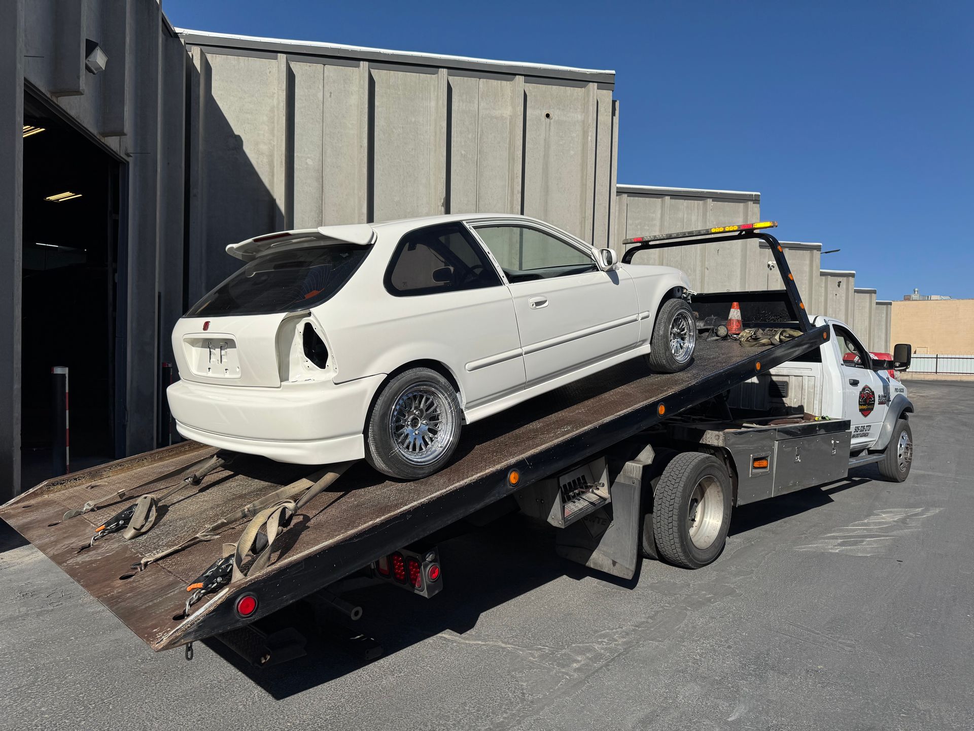 White hatchback on a flatbed tow truck beside a warehouse in an industrial lot.