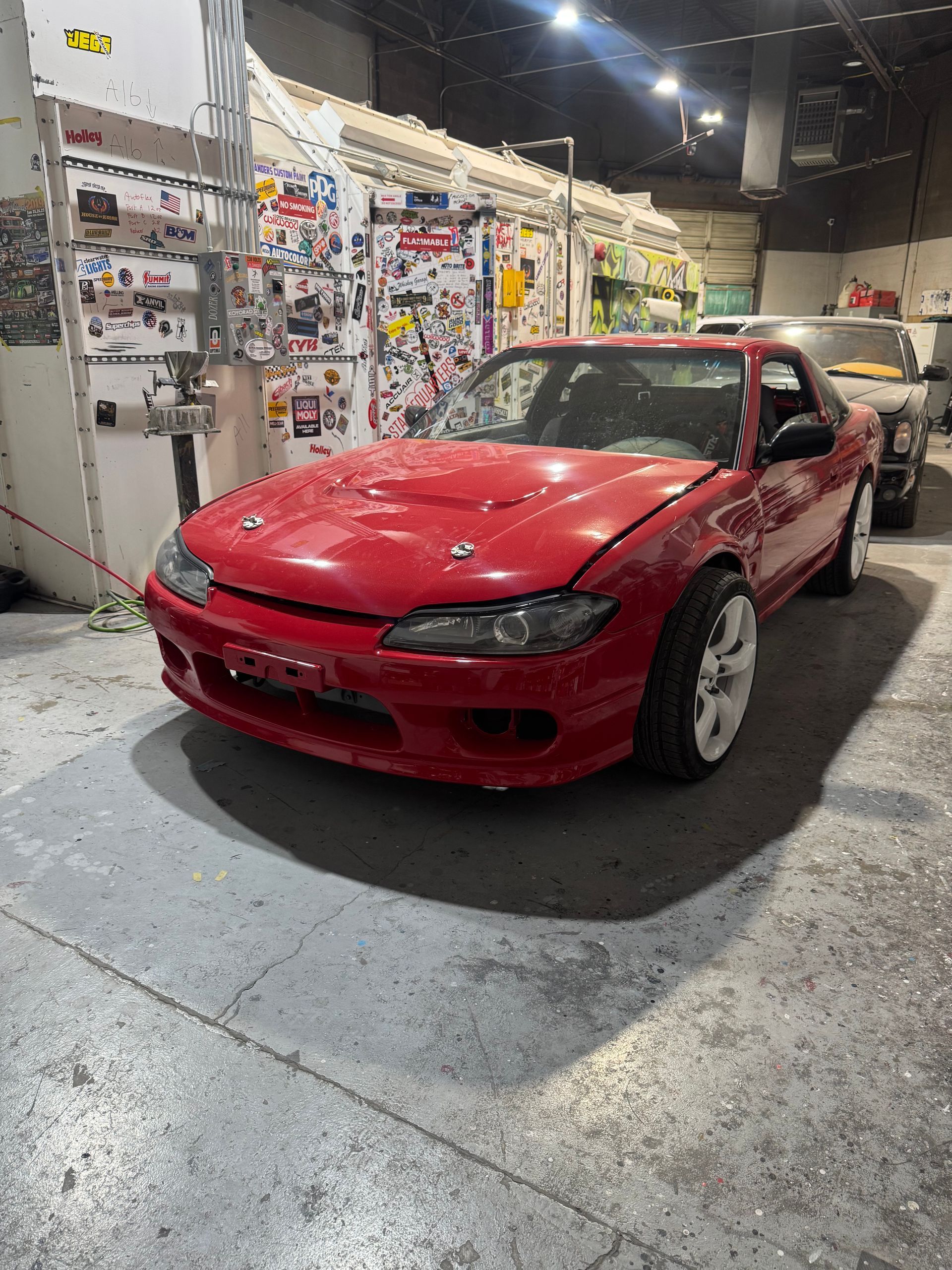 Red sports coupe in an indoor garage, red hood raised and front bumper removed, with wall displays behind it