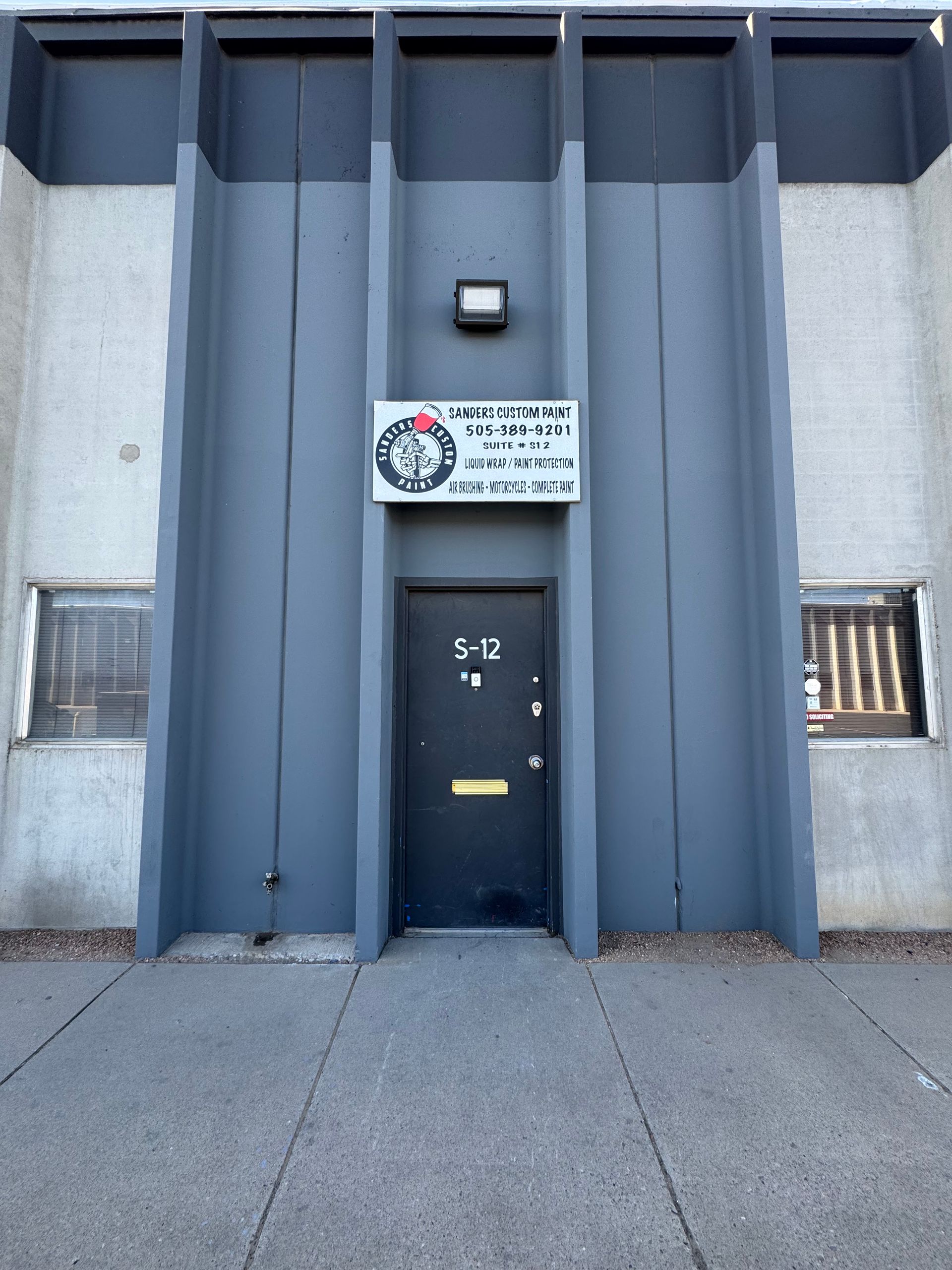 Gray building entrance with a centered door and sign above it, flanked by windows.