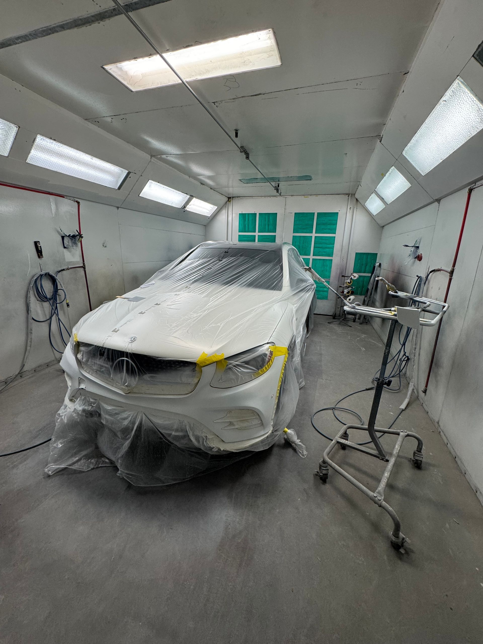 Car covered in plastic inside a bright auto body paint booth, with tools and equipment nearby.