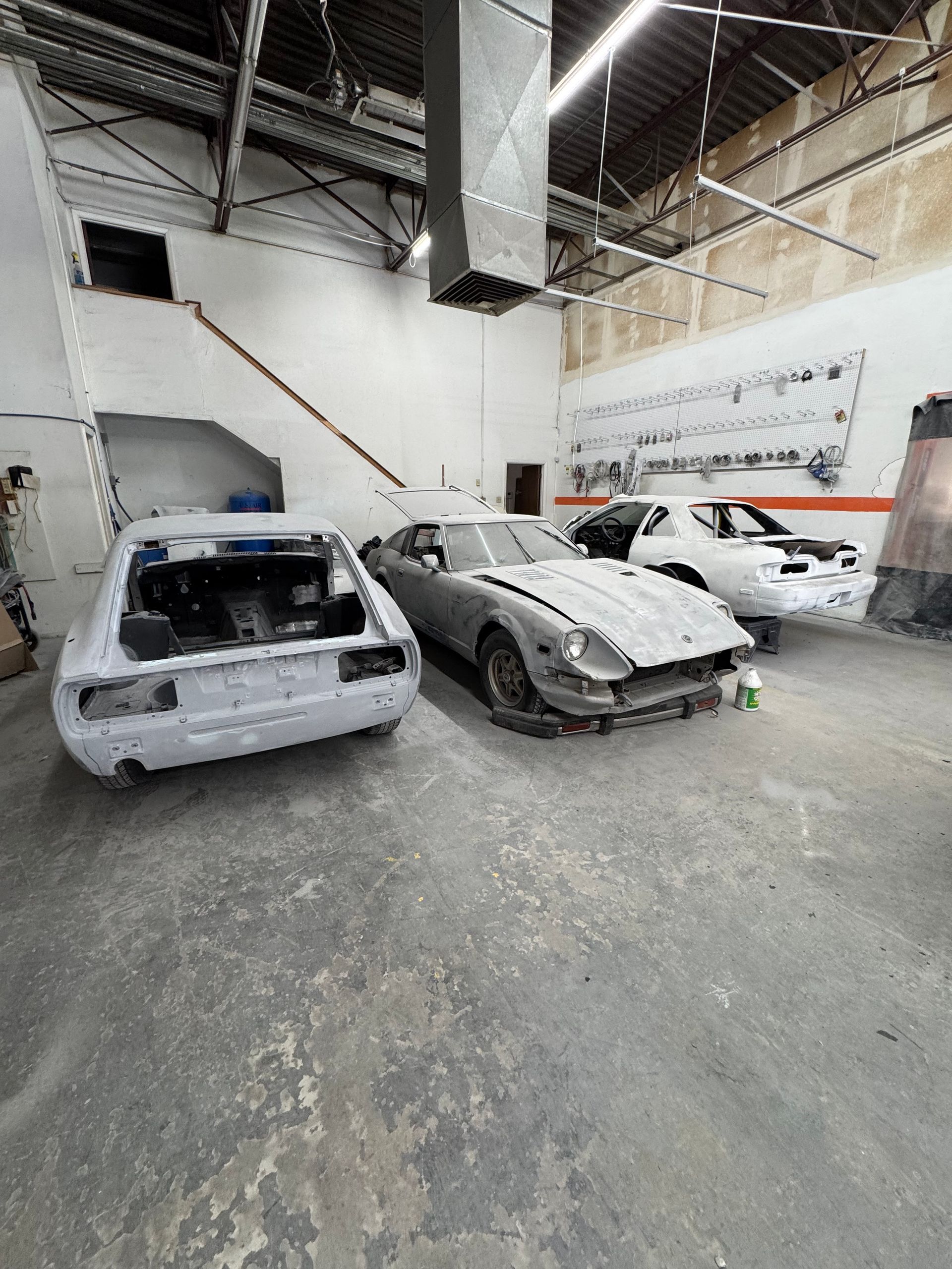 Three damaged cars in an auto body shop with a high ceiling and industrial lighting.