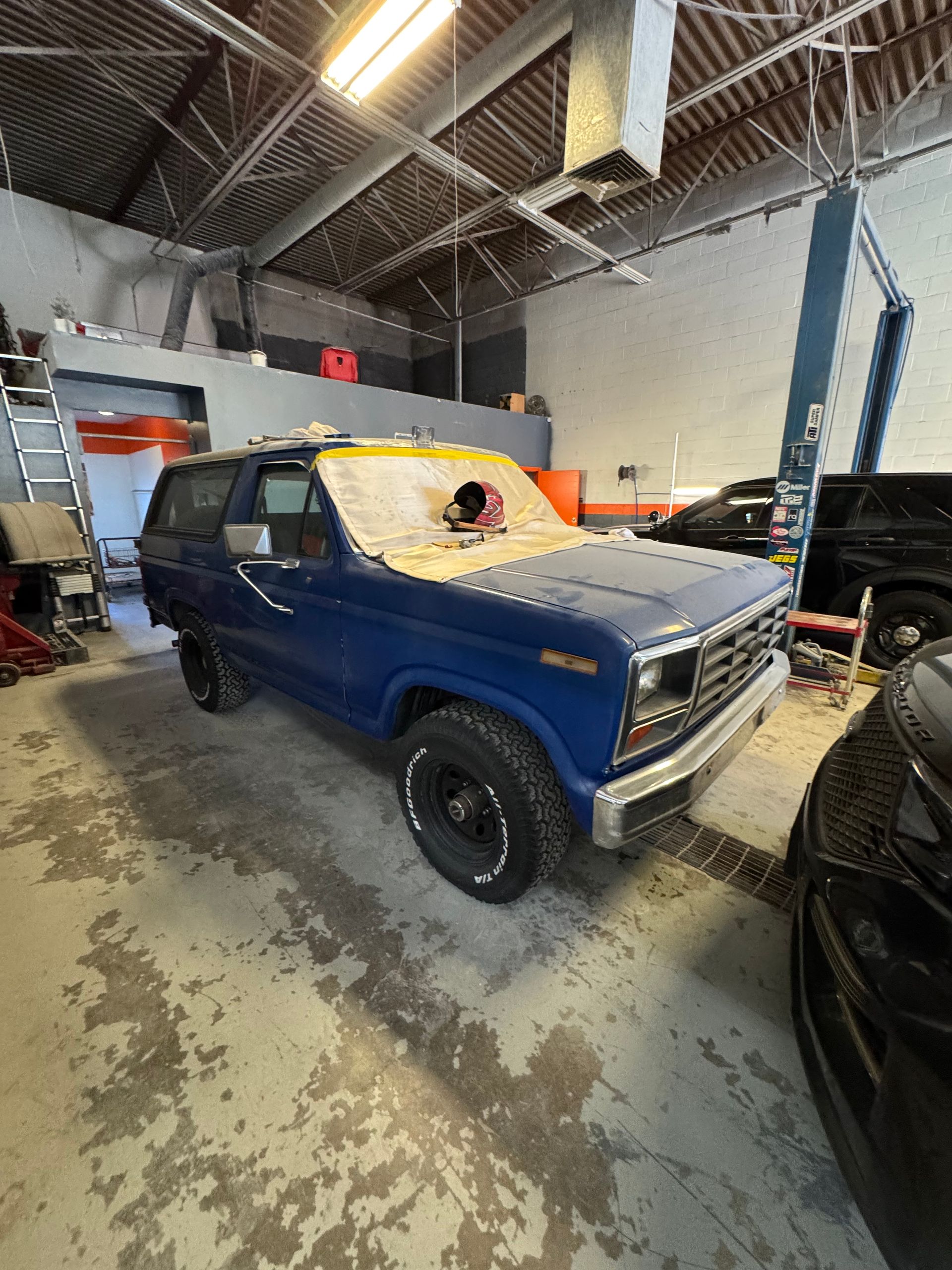 Blue vintage SUV in an auto shop, with an orange tarp over the roof and windows.