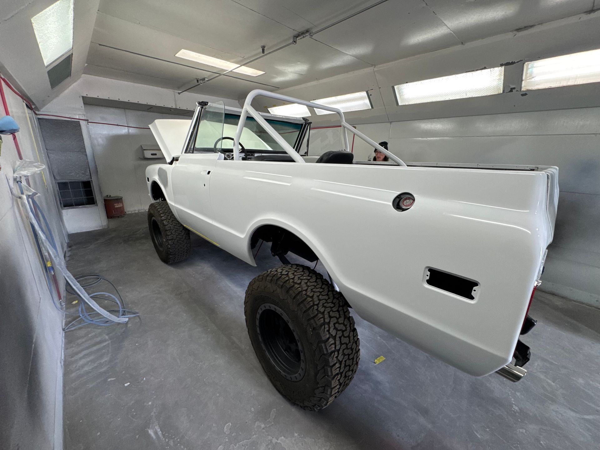 White off-road truck body in a paint booth with large black tires and a bare frame.