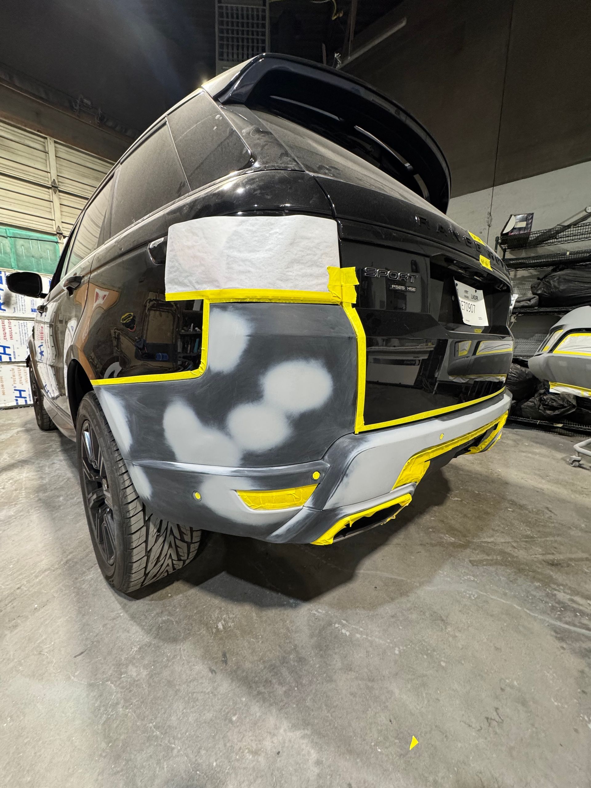 Damaged black SUV bumper in a repair shop, with gray primer patches and yellow outline markings
