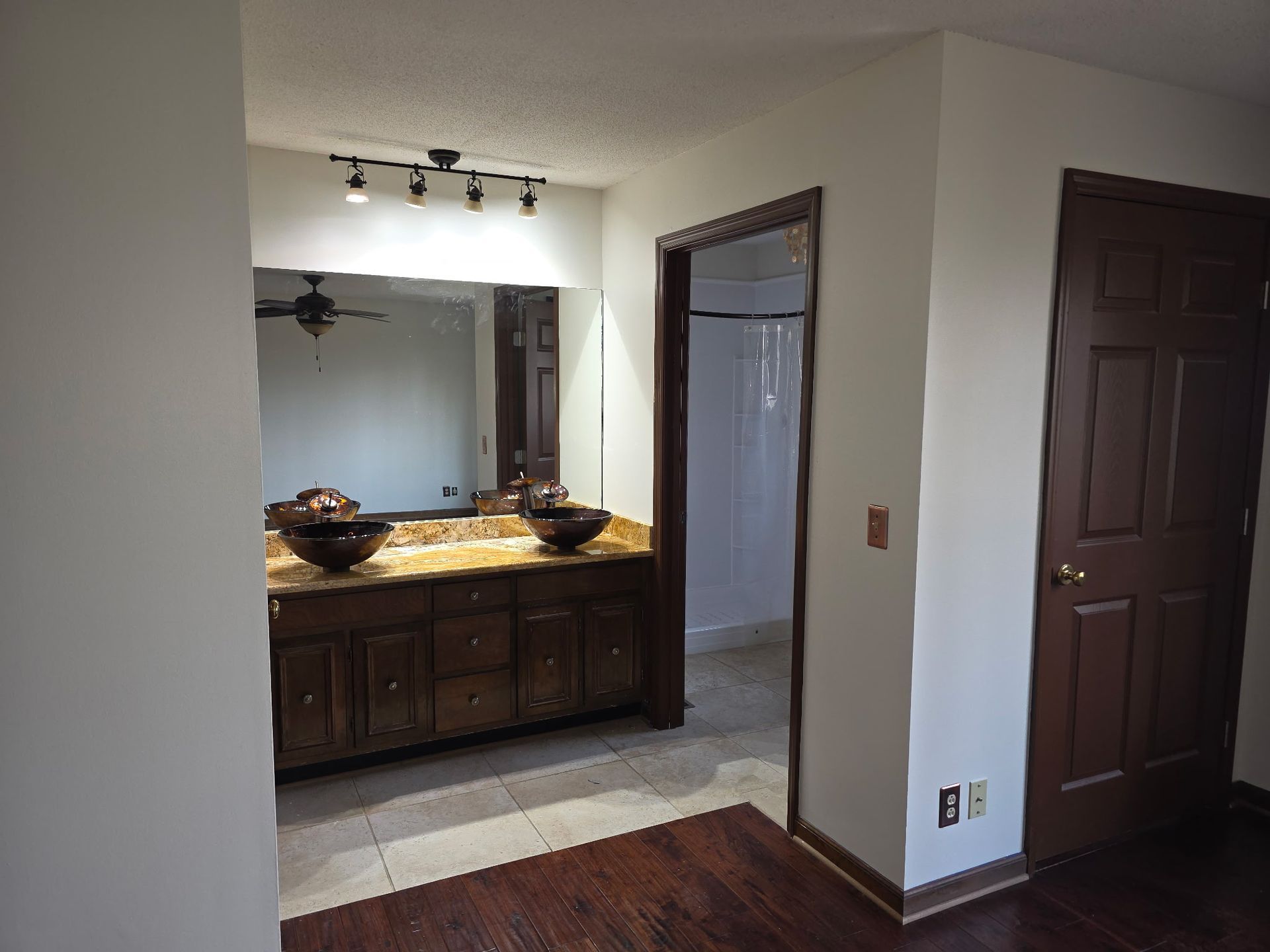 A bathroom vanity with dark wood cabinets, a tan countertop, two vessel sinks, a large mirror, and a dark brown door.