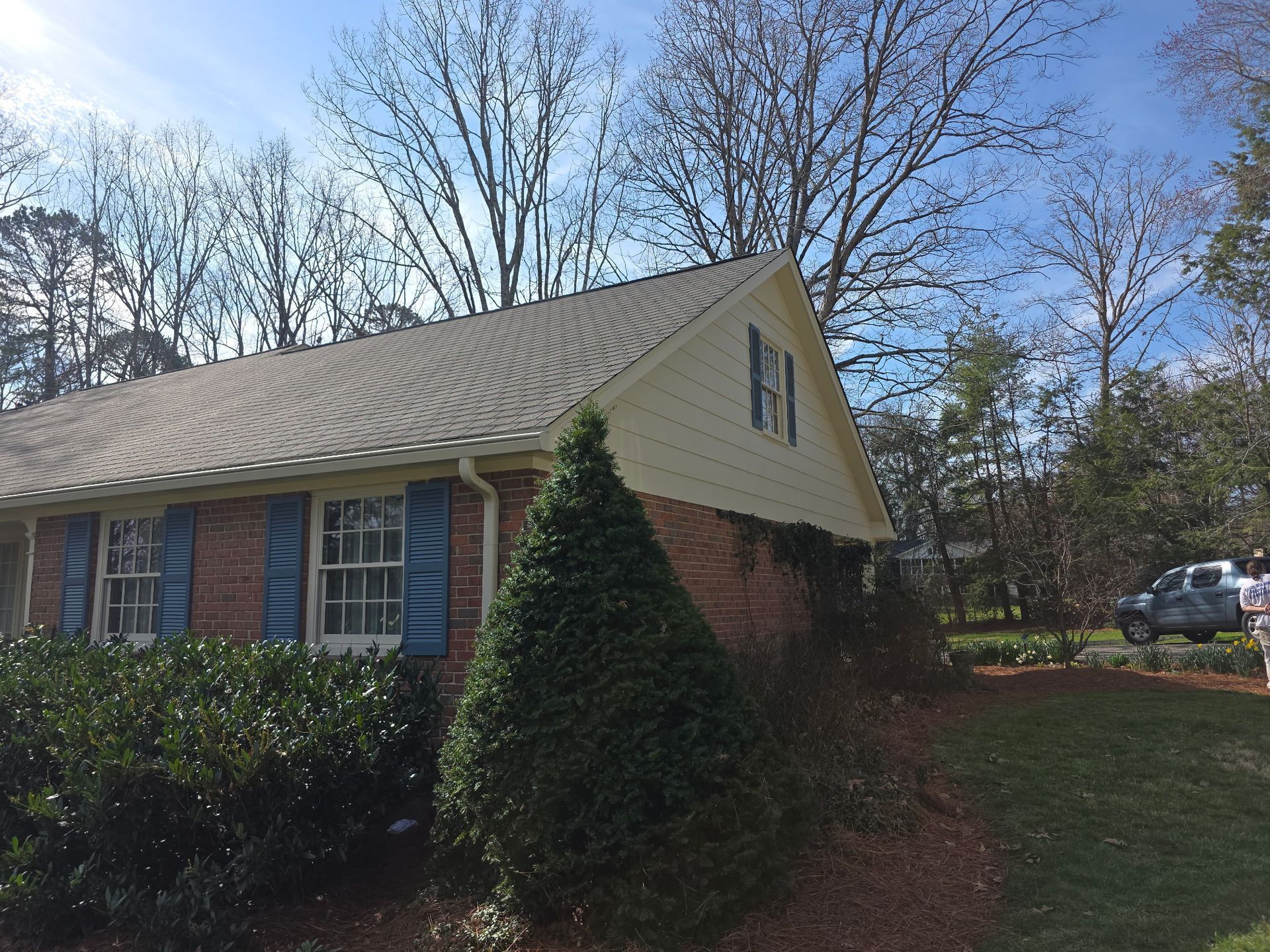 A light brick house with blue shutters and cream siding under a gable roof, surrounded by trees and landscaping.