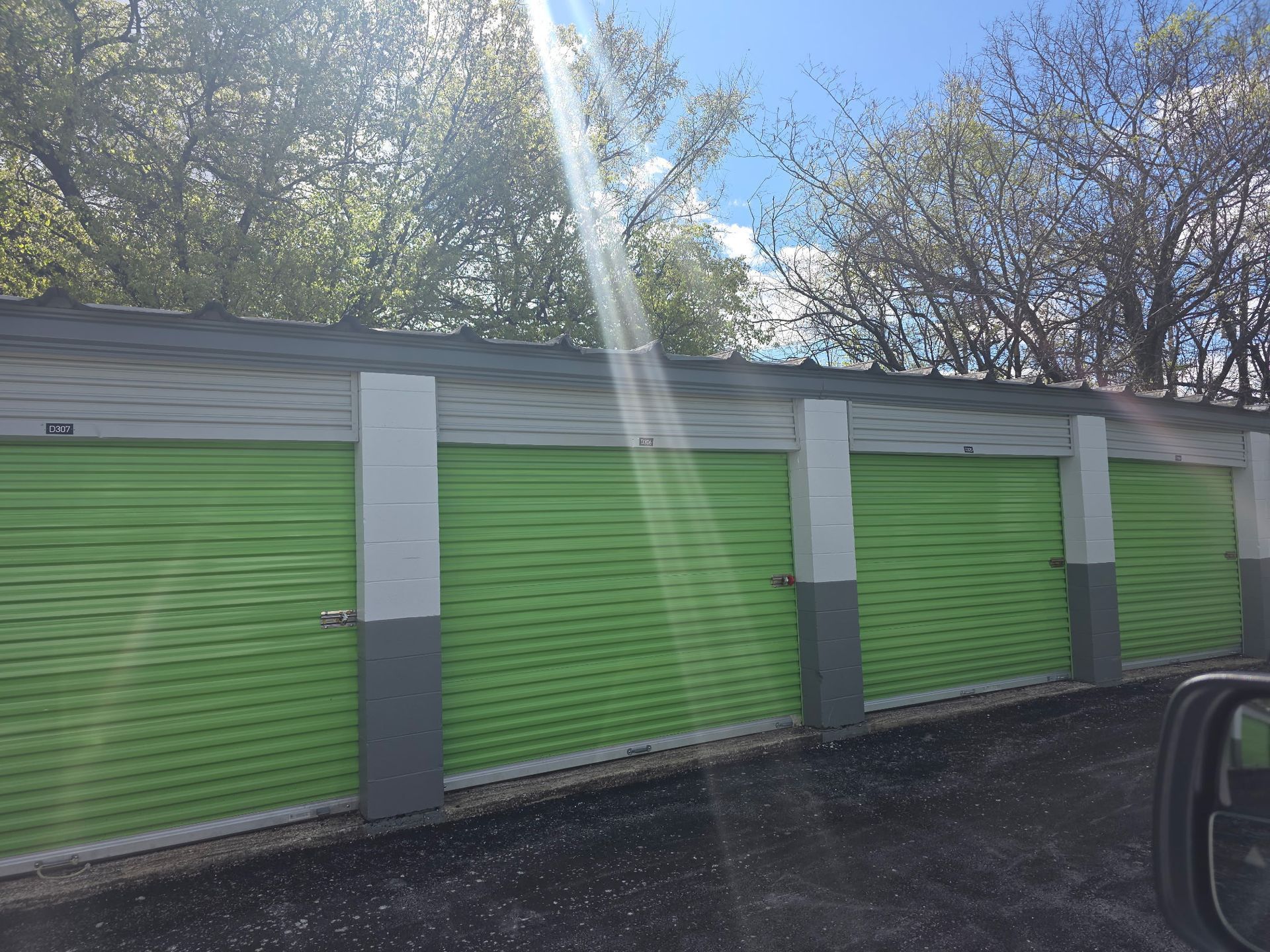 A row of storage units with lime green rolling doors separated by grey pillars under trees on a sunny day.