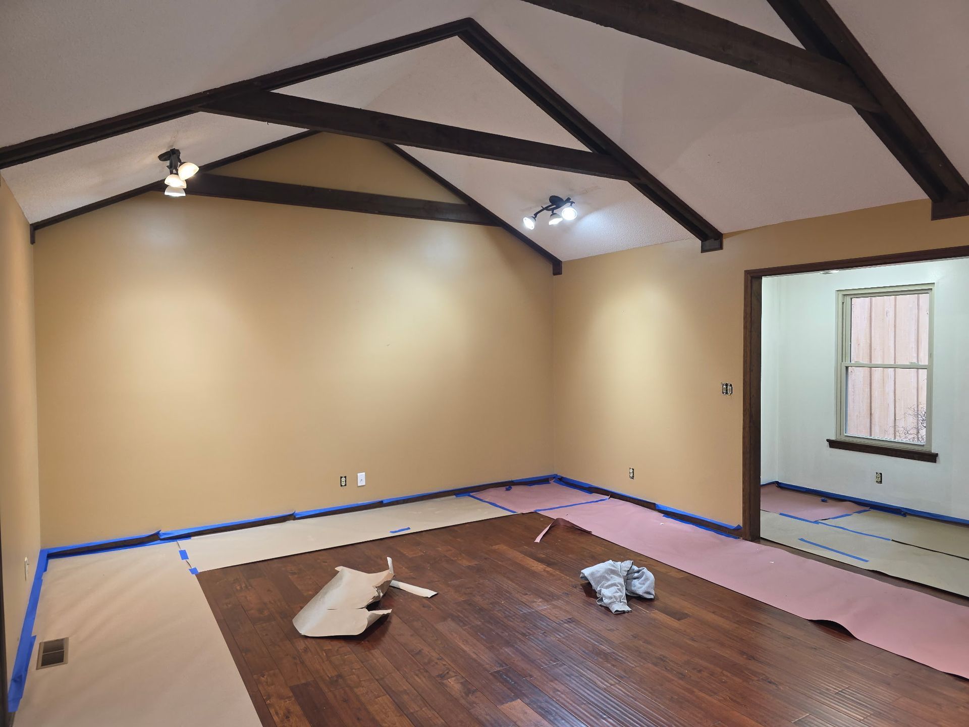 An interior room under renovation with tan walls, dark wood floors, vaulted ceilings with beams, and protective paper.