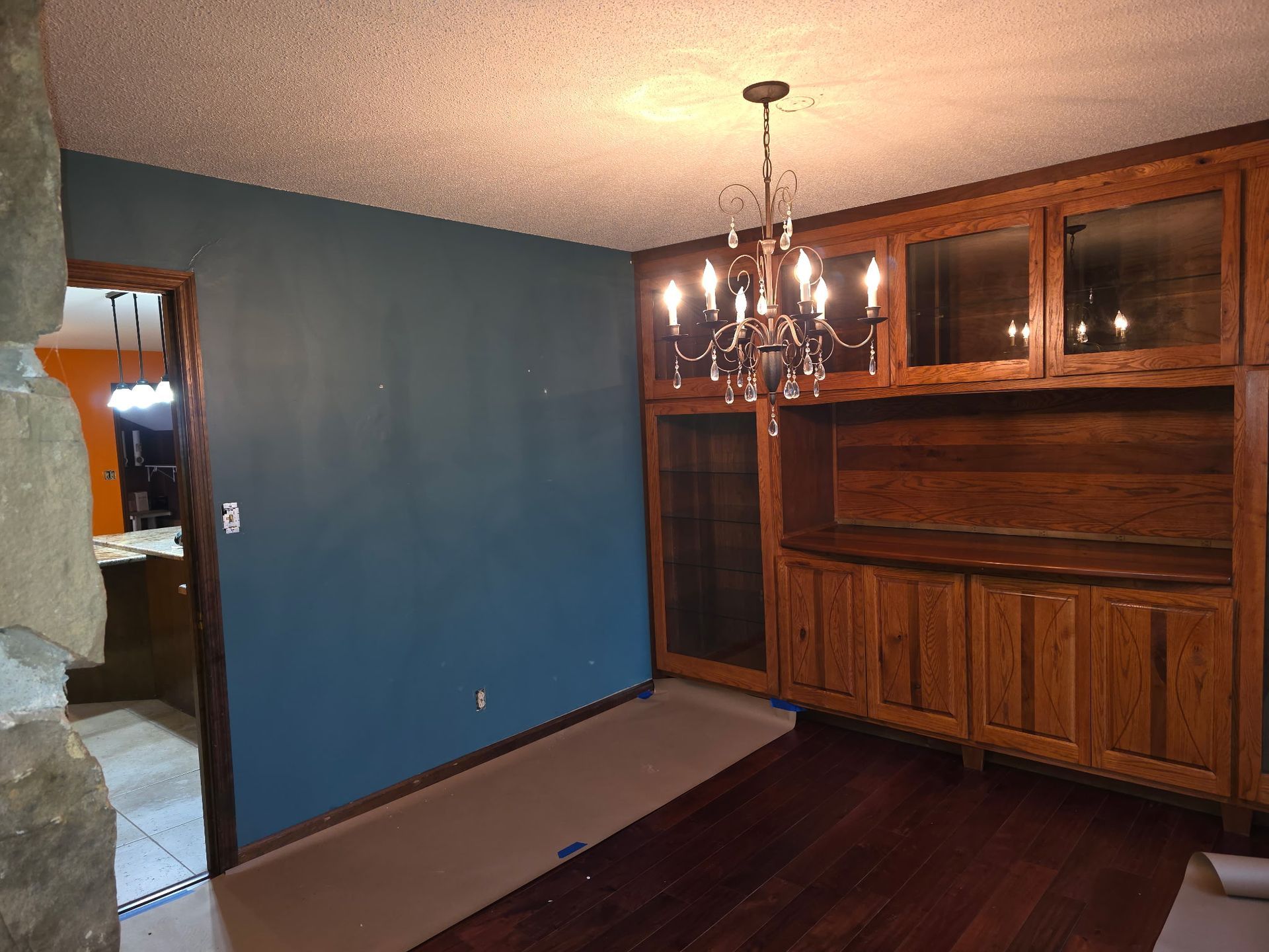 A dining room with dark teal walls, a rustic chandelier, wood cabinetry, and a stone pillar in the foreground.