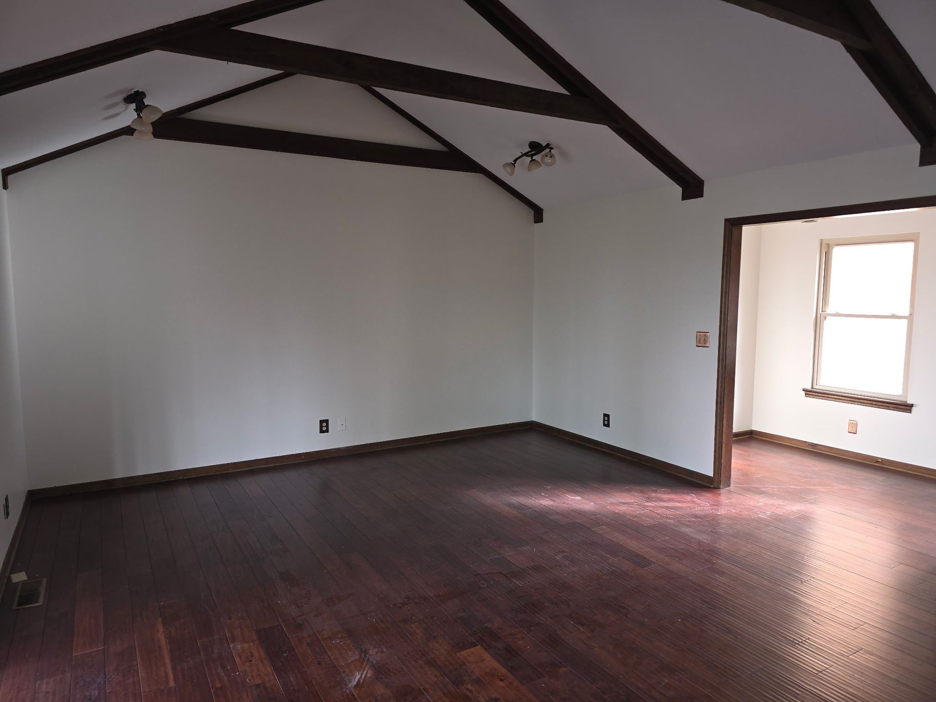 A spacious room with white walls, dark wooden floors, exposed ceiling beams, and an open doorway leading to a lit room.