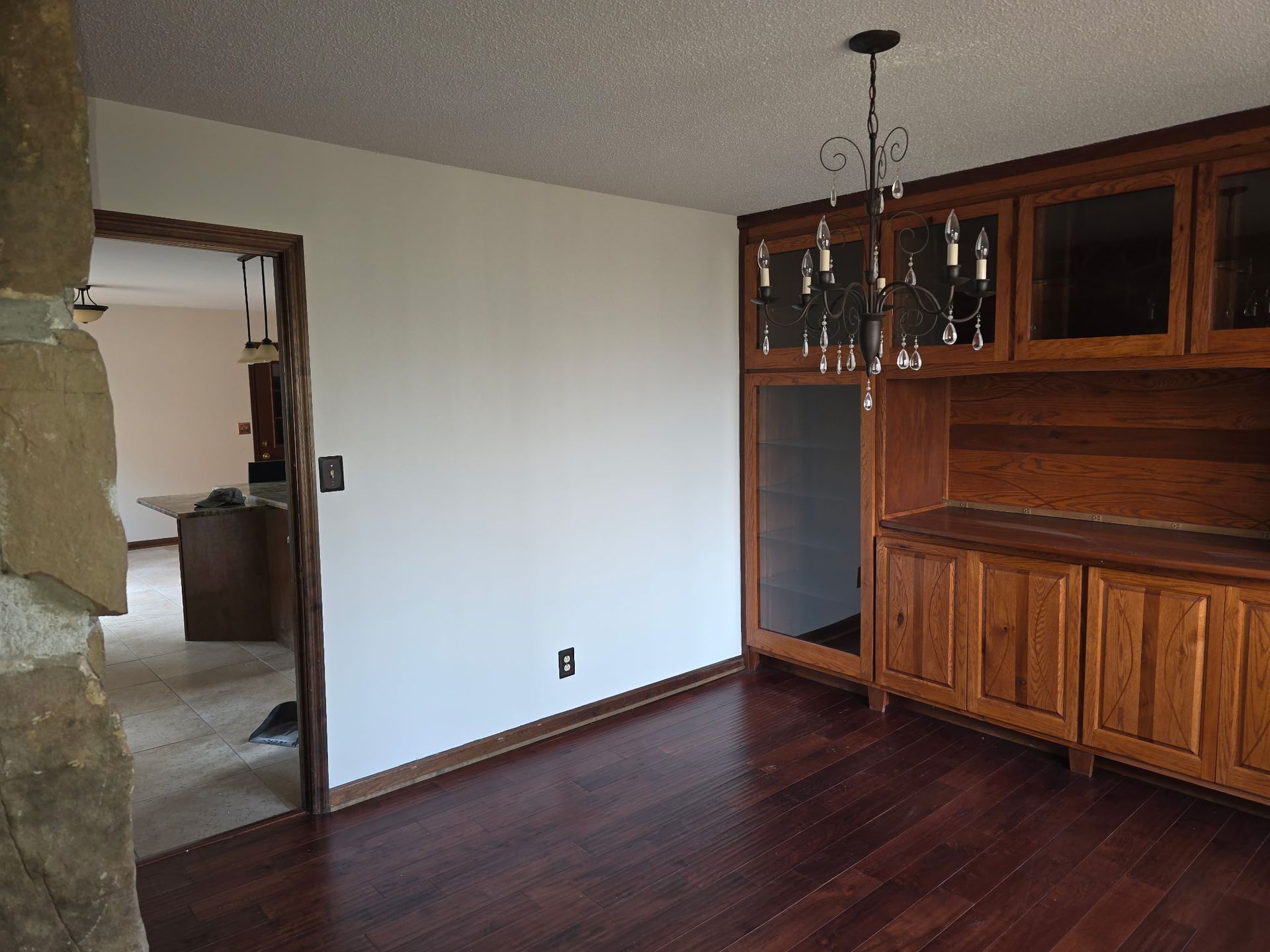 A dining room with dark wood flooring, a built-in wooden cabinet, and a chandelier, viewed through a stone archway.