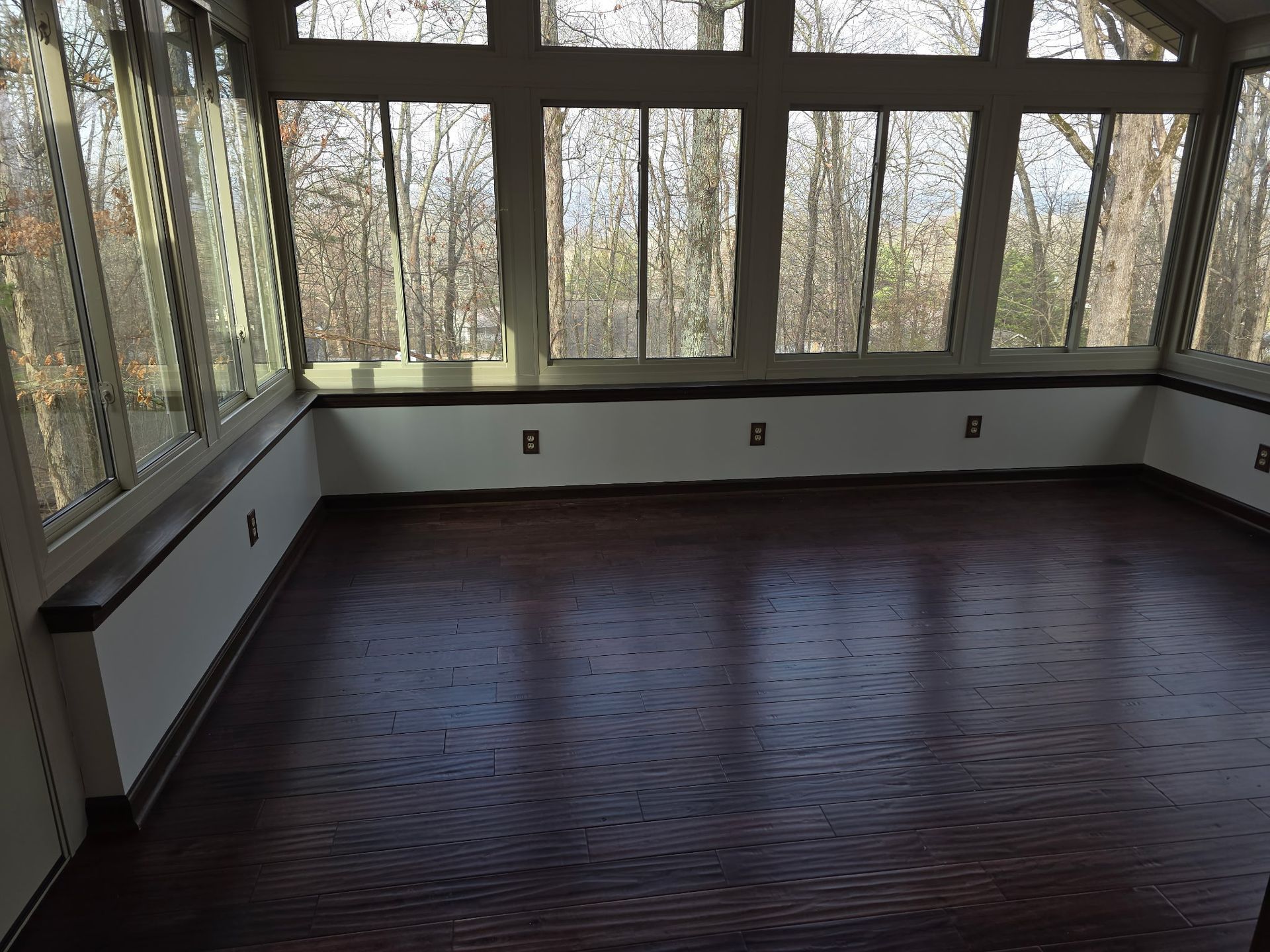 A bright sunroom with dark wood floors, white walls, and a large bank of windows overlooking a wooded area.