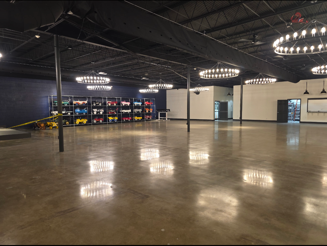 Spacious empty indoor arena with glossy floor, hanging circular lights, and shelves of colorful equipment at left