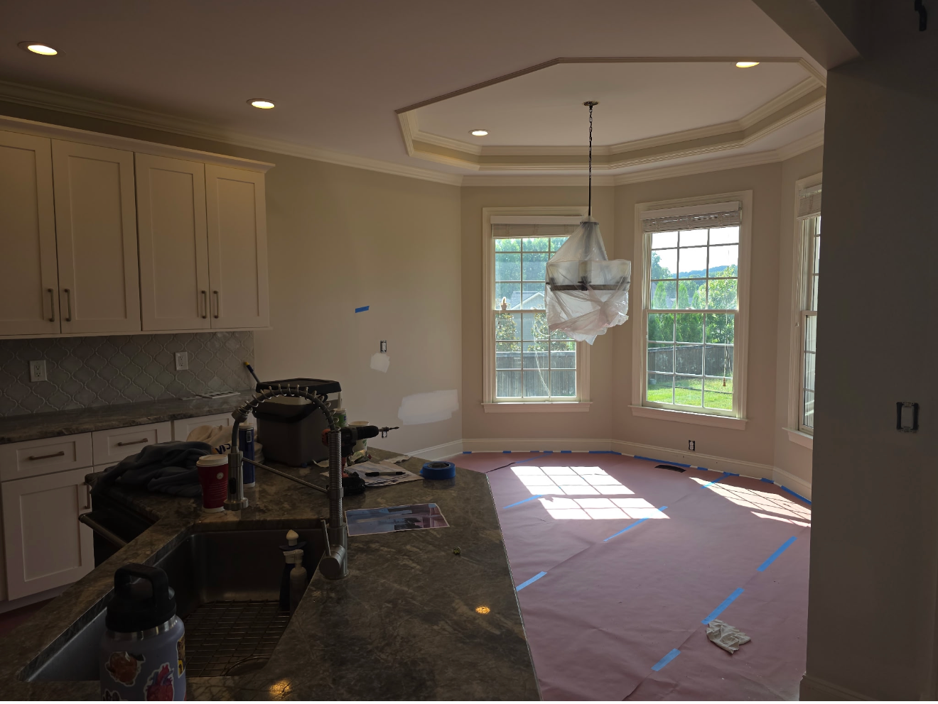 Kitchen and dining area under renovation, with cabinets, tools, and taped floor near bay windows.