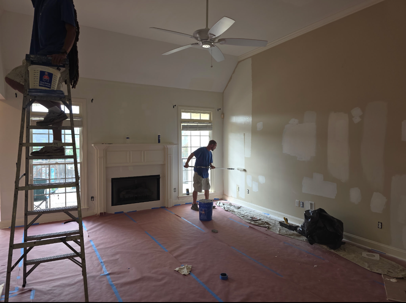 Painter on ladder in a room with covered floor, fireplace, and patchy drywall repairs.