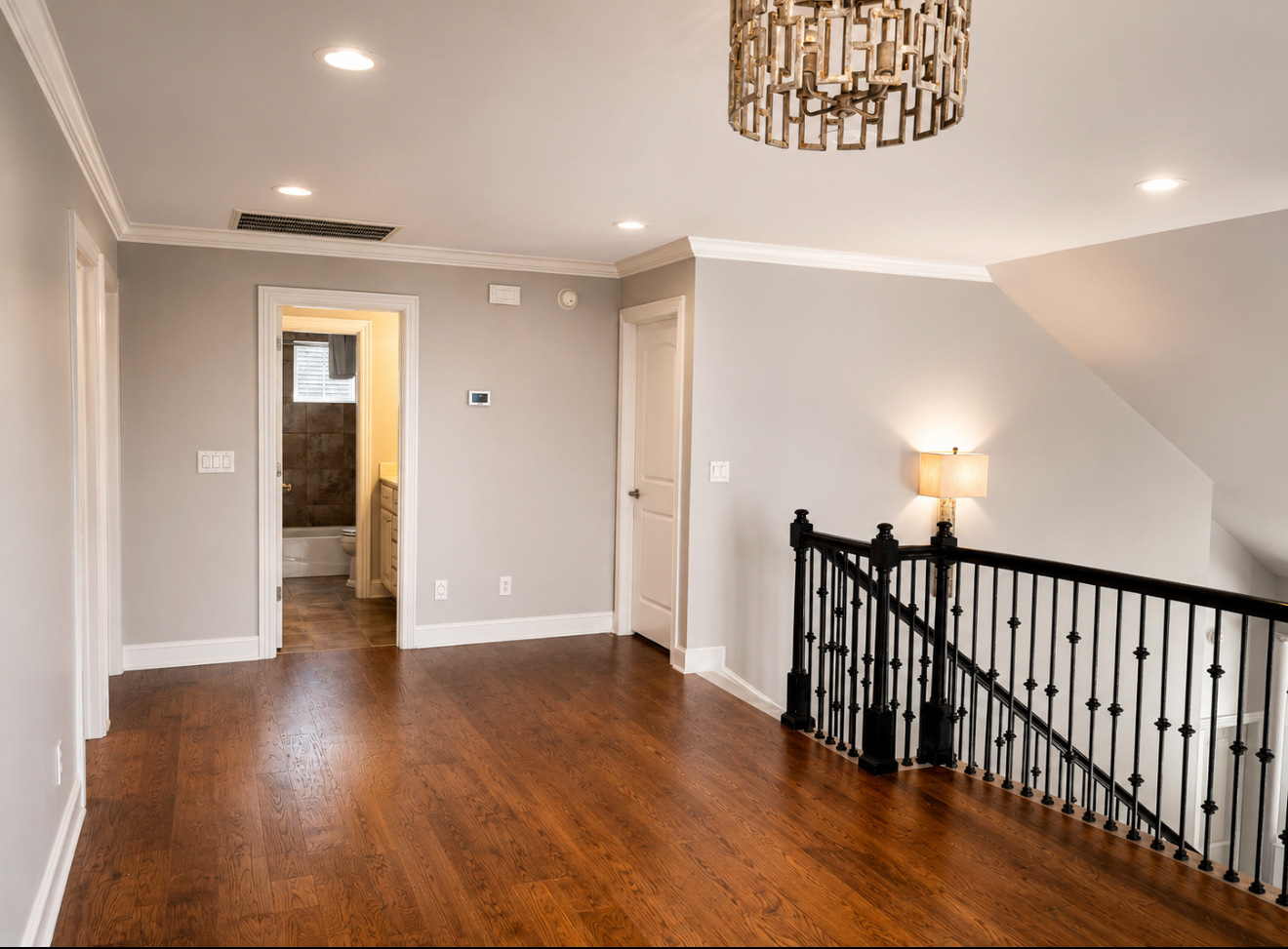 Empty upstairs hallway with hardwood floors, gray walls, black stair railing, and a doorway to a bathroom.