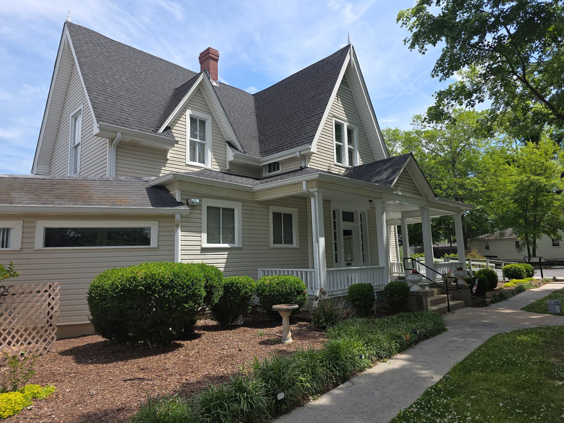 Victorian-style gray house with steep roof, front porch, and landscaped yard on a sunny day