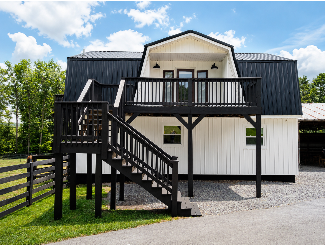 White house with black roof, front stairs, and a small balcony on a gravel driveway beside a grassy yard.
