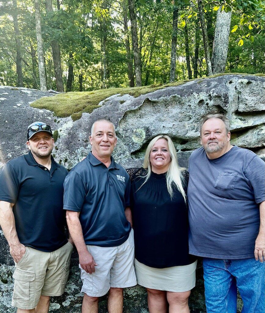 Four people standing in front of a large rock, trees in the background.