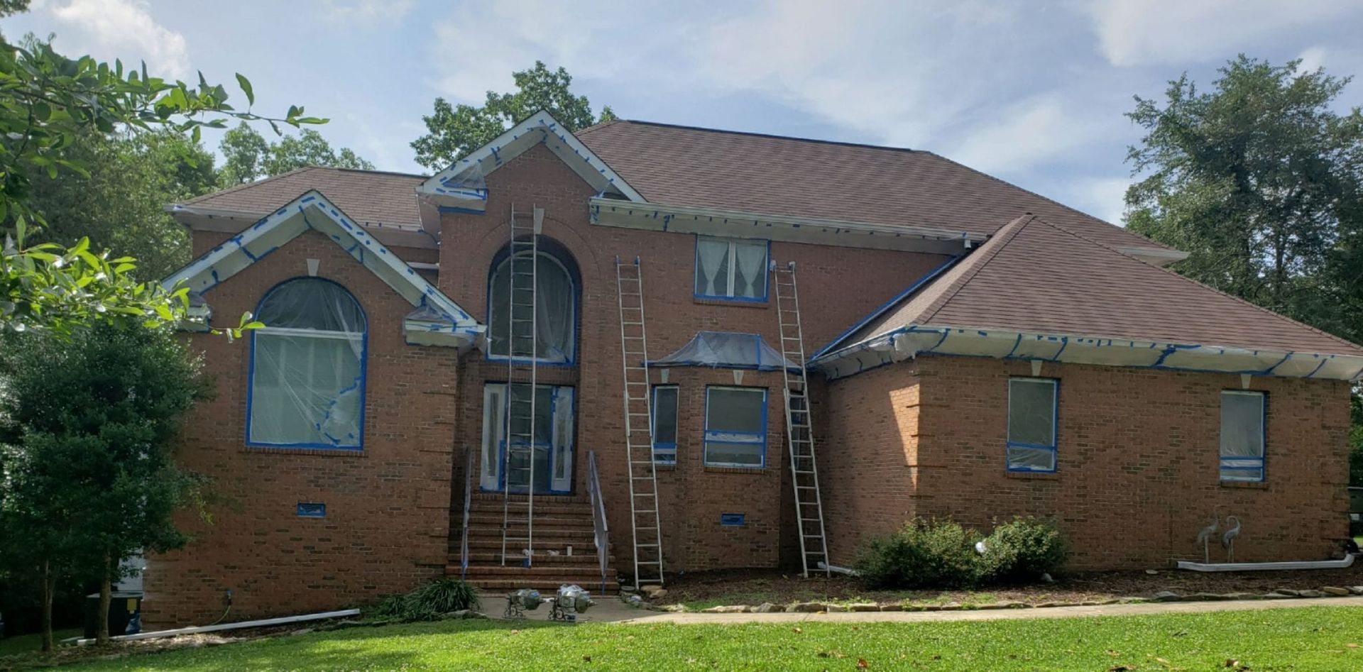 A brick house under construction with ladders, surrounded by trees and green grass.