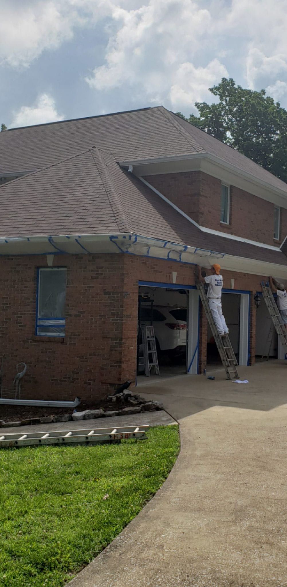 Brick house with a brown roof, workers on ladders near the garage, sunny day.