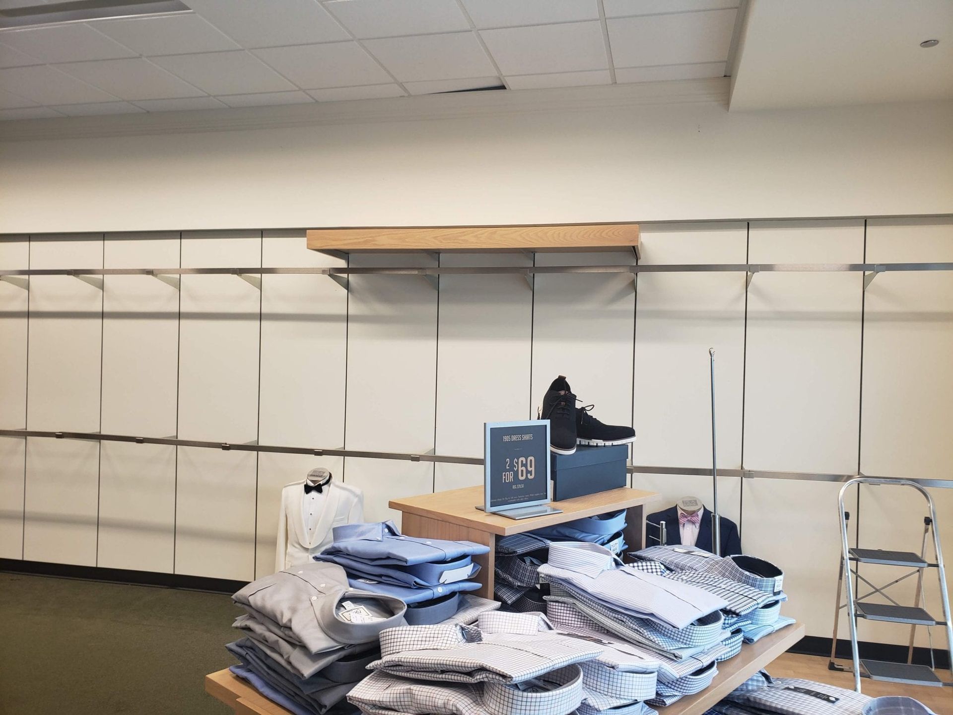 Clothing store display with shirts and shoes. Beige walls with shelving and a ladder.