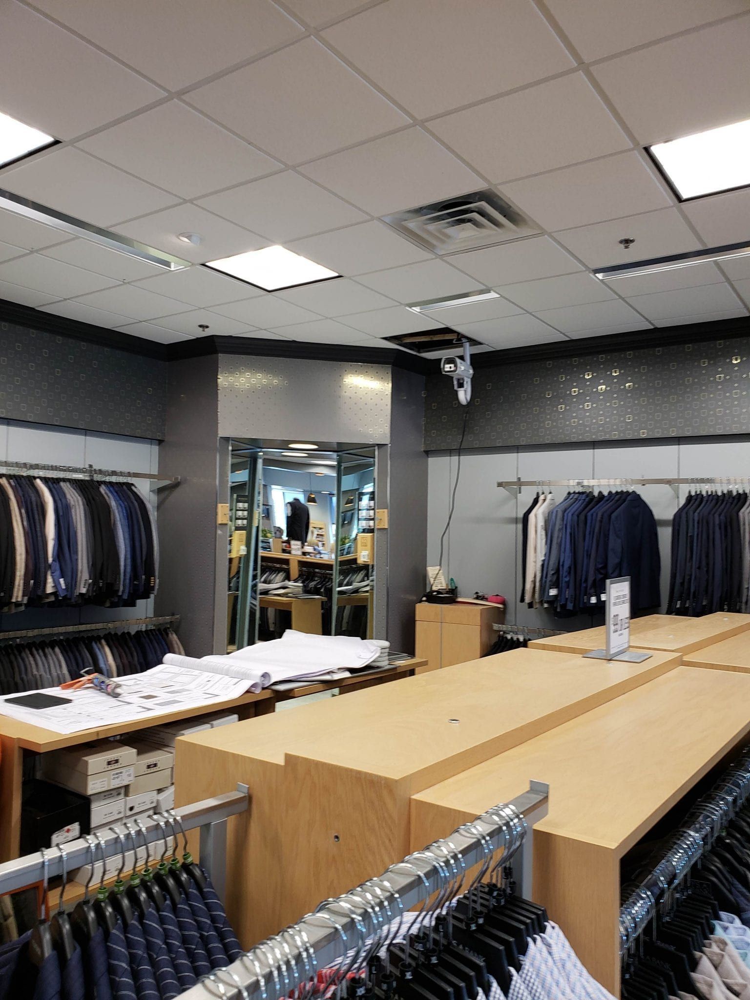 Clothing store interior with racks of suits and shirts. Work tables and a central pillar.