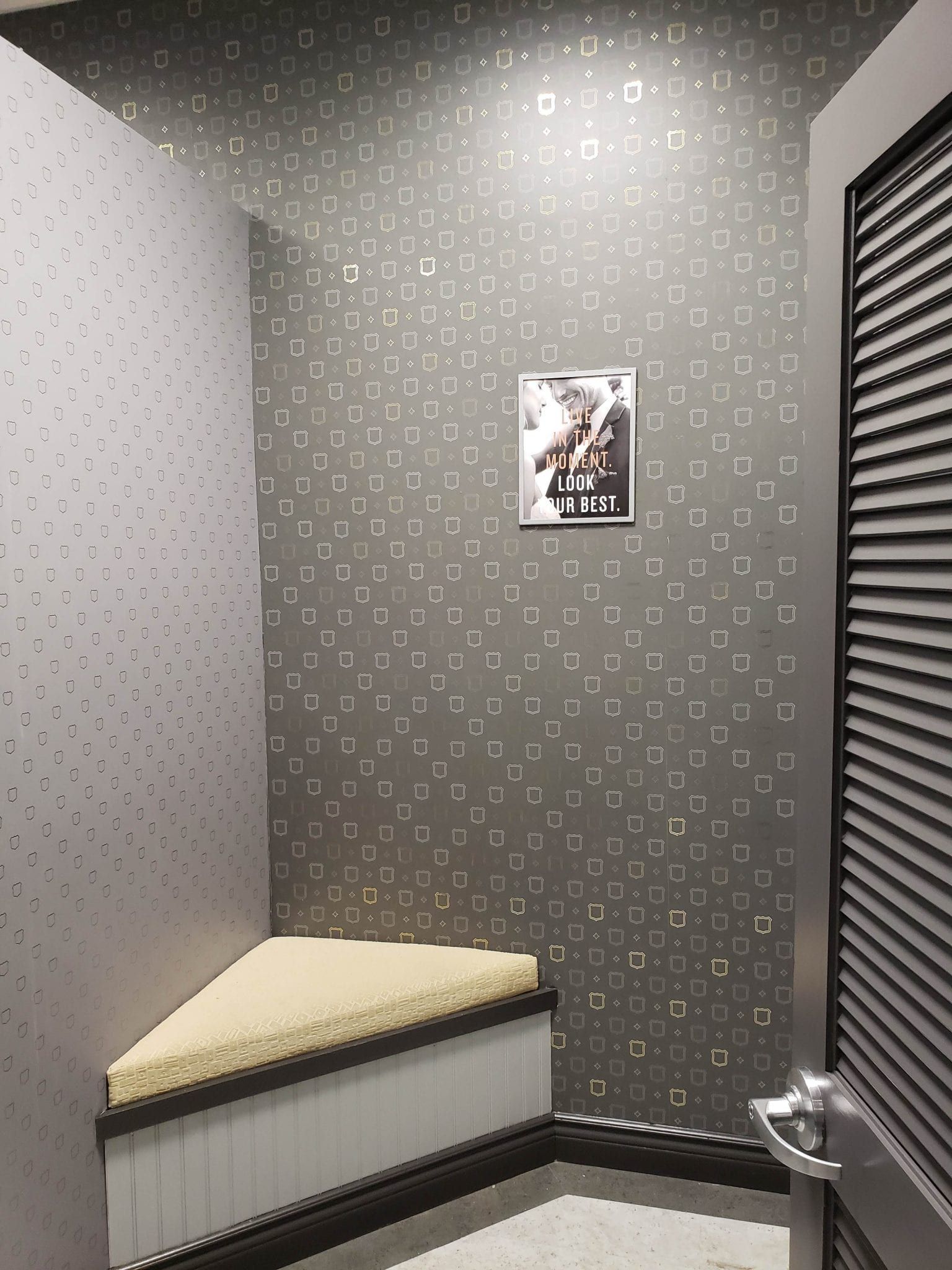 A small, gray, geometric-patterned room with a built-in bench and a framed image. A louvered door is on the right.