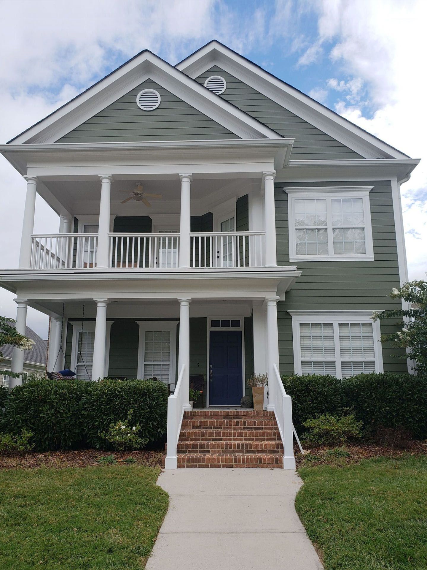 Two-story green house with white trim, a covered porch, and a brick walkway leading to the front door.